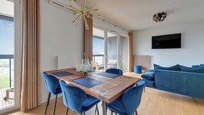 A modern living room with a wooden dining table, blue chairs, a blue sofa, and a mounted TV. - Horizontal Sea & City view | Gdynia | 3 bedrooms (Gdynia)