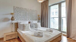 En pænt redt seng med hvide sengetøj og puder, et natbord med en lampe, og et stort vindue med udsigt til bygninger.