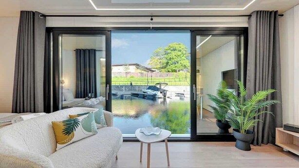 A modern living room with a sofa, a small table, and a large sliding glass door leading to a courtyard with a view of a canal and boats. - Houseboat Aurora | Old Town | Romantic | Sauna (Gdańsk)