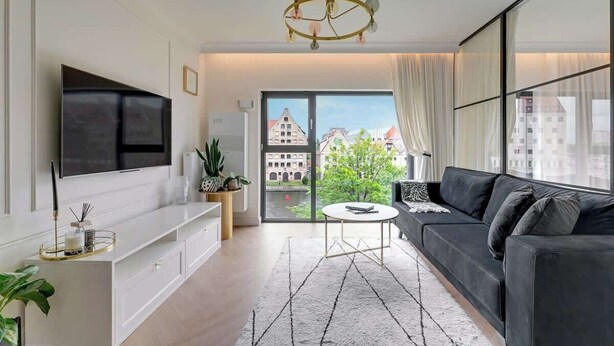 A modern living room with a flat-screen TV, a black sofa, a round coffee table, and a chandelier. - Rezydencja Wintera 1 | River View (Gdańsk)