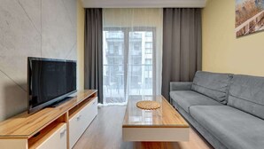 A modern living room with a flat-screen TV, a grey sofa, a wooden coffee table, and a large window with curtains. - Zajezdnia Wrzeszcz 9 | Balkony | Parking (Gdańsk)