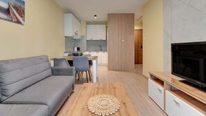 A modern living room with a sofa, TV, and a dining area with chairs and a table. - Zajezdnia Wrzeszcz 9 | Balkony | Parking (Gdańsk)
