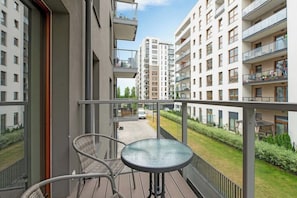Un balcon doté d’une table en verre et de deux fauteuils en osier, donnant sur un quartier résidentiel avec des immeubles modernes.