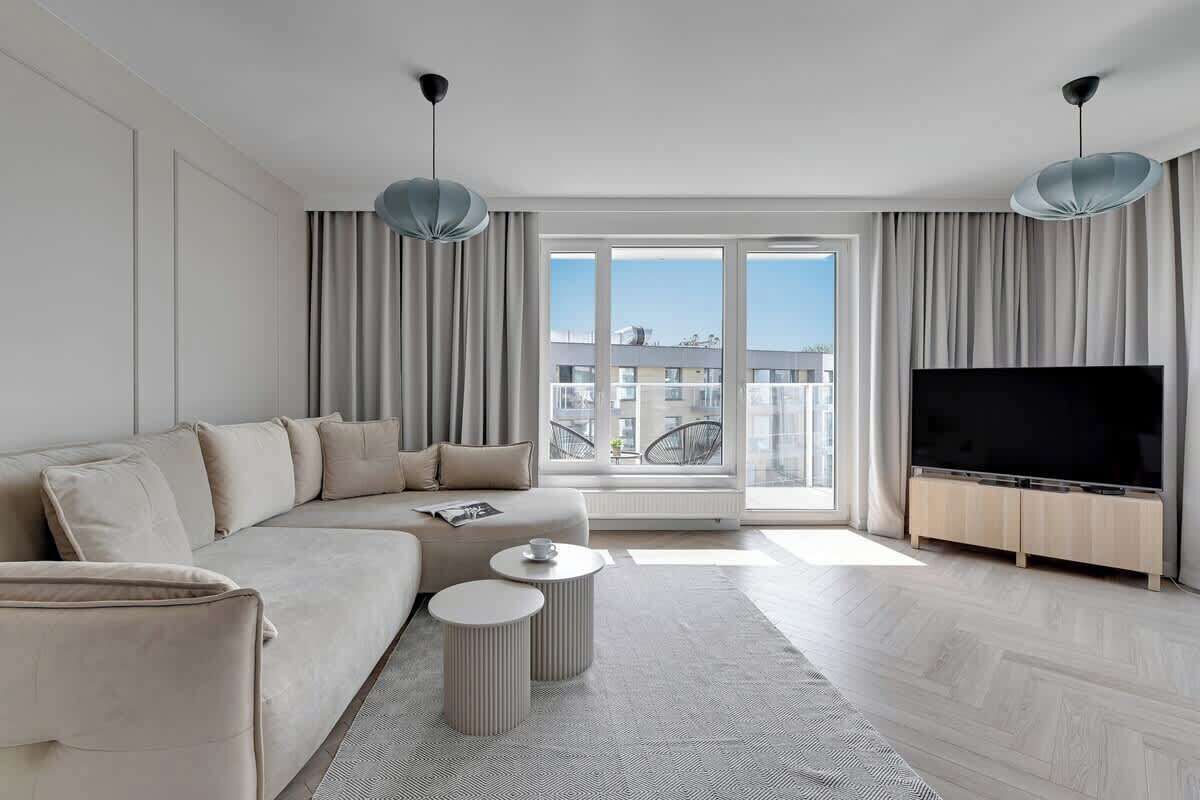 Un salon moderne avec un canapé, deux tables d’appoint, un téléviseur à écran plat et une grande fenêtre avec des rideaux.