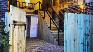 A covered outdoor area with a wooden staircase leading to a second-floor balcony, surrounded by a brick wall and a wooden fence. - The Little Building | Historic Hocking Hills (Logan)