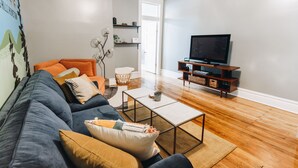 A living room with a sofa, coffee table, TV stand, and a door leading to another room. - The Little Building | Historic Hocking Hills (Logan)