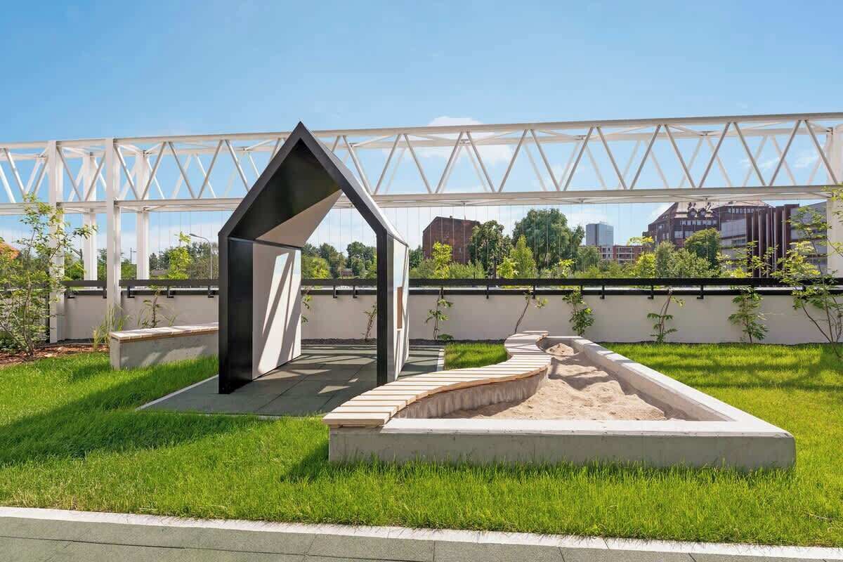 A modern outdoor space with a geometric structure, a concrete planter, and a metal bridge in the background.