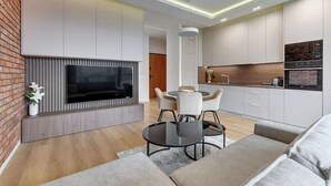 Uma sala de estar moderna com televisão de tela plana, um espaço de refeições com mesa redonda e cadeiras, e uma cozinha com armários brancos e painel de parede de tijolos.