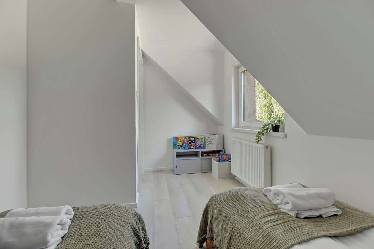 Un dormitorio con dos camas, una ventana, un radiador y una pequeña estantería con objetos.