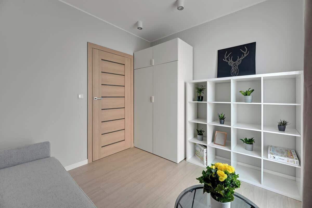 A modern living room with a grey sofa, a wooden door, white cabinets, and a white shelving unit with plants.