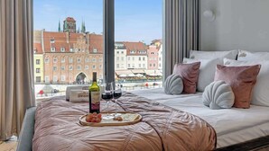Habitación de hotel con un ventanal grande, cama con cubrecama rosa y una bandeja con vino y aperitivos.