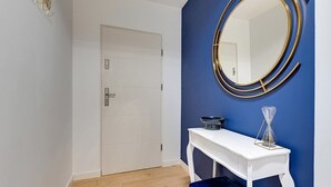 Ein Zimmer mit einer weißen Tür, einem weißen Beistelltisch mit einer blauen Bank, einem runden Spiegel mit goldener Rahmung und einer kleinen Sanduhr auf dem Tisch.