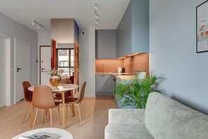Ein modernes Wohnzimmer mit Sofa, Esstisch mit Stühlen, Küchenzeile und einer großen Pflanze.