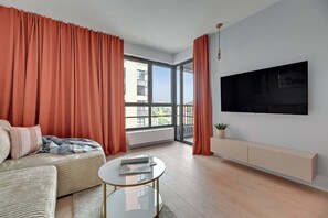 Ein modernes Wohnzimmer mit Sofa, Couchtisch, Wandfernseher und großen Fenstern mit Vorhängen.