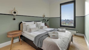 Een net opgemaakt bed met kussens en een deken, een nachtkastje, een wandklok en een raam met uitzicht op de stad.