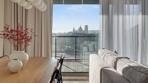 Een moderne woonkamer met een bank, een eettafel met stoelen, een vaas met rode bloemen en een uitzicht op een stadsgezicht door grote ramen.