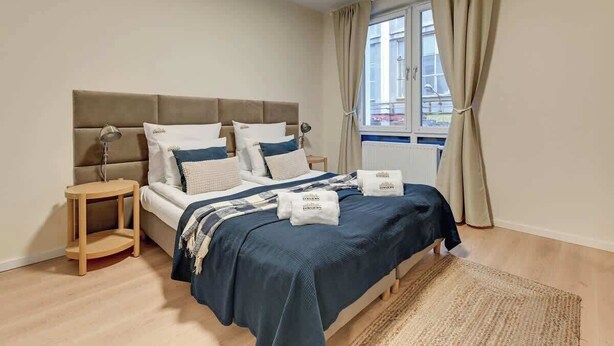 Un dormitorio con cama, mesitas de noche, una ventana con cortinas y vistas a edificios.