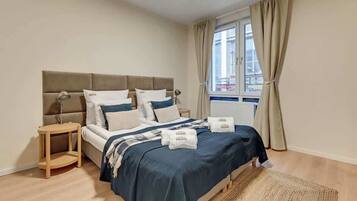Ein Schlafzimmer mit Bett, Nachttischen, einem Fenster mit Vorhängen und Blick auf Gebäude.