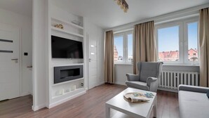 Ein modernes Wohnzimmer mit Flachbildfernseher, grauem Sofa, weißem Couchtisch und großen Fenstern mit Vorhängen.