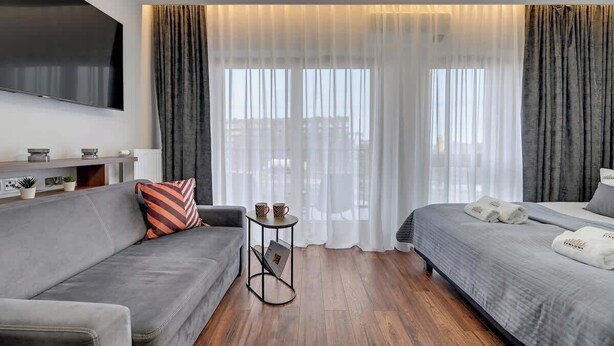 Ein modernes Hotelzimmer mit grauem Sofa, einem Bett mit grauer Bettwäsche, einem kleinen Beistelltisch mit einer Zeitschrift und einem an der Wand befestigten Fernseher.