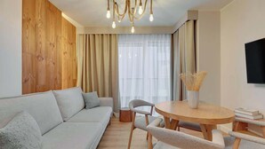 Een moderne woonkamer met een bank, een ronde houten tafel, stoelen en een aan de muur bevestigde televisie.
