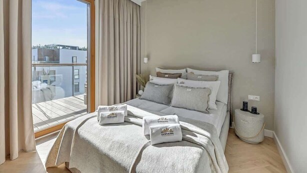 Ein Schlafzimmer mit Bett, Kissen, einem Nachttisch, einem Fenster mit Vorhängen und Blick auf Gebäude.