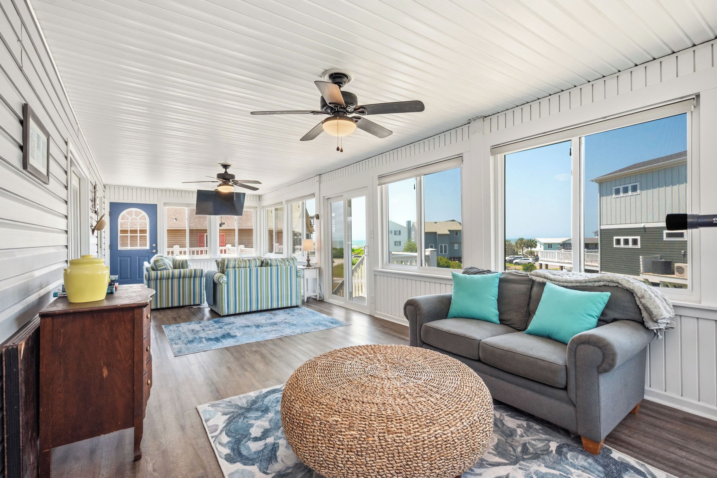 A spacious room with a large window, a ceiling fan, a gray sofa with teal cushions, and a wicker ottoman.