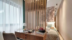 Moderni hotellihuone, jossa on työpöytä, tuoli, sänky ja suuri, abstrakti taulu seinällä.