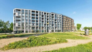 Un edificio moderno de varios pisos con balcones, un área verde al frente y un cielo despejado.