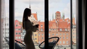 A person standing by a window overlooking a cityscape with buildings and a church. - Deo Plaza 306 | View of the river and the Old Town (Gdańsk)
