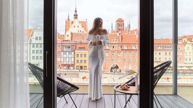 A room with a large window overlooking a cityscape, a table with a basket of fruit, and two chairs. - Deo Plaza 306 | View of the river and the Old Town (Gdańsk)