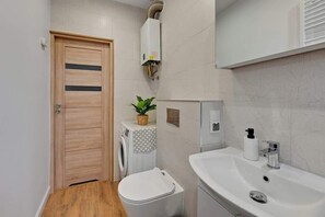 Une petite salle de bain avec une porte en bois, un lave-linge, des toilettes, un lavabo avec un distributeur de savon et une plante en pot.