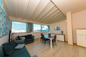 En moderne stue med sofa, spisebord og stole, et køkkenområde med bordplade og skabe, samt store vinduer med gardiner.