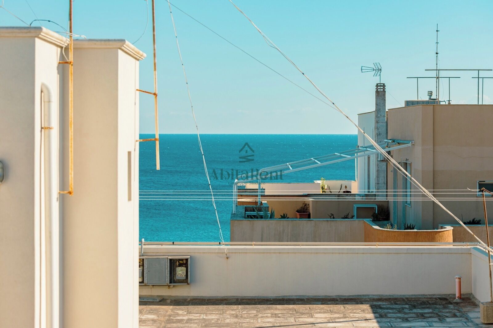 A rooftop terrace with a view of the sea, a clear sky, and a building with antennas.
