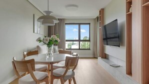 Een moderne eetruimte met een ronde tafel, zes stoelen, een vaas met bloemen, een groot raam met uitzicht en een tv aan de muur.