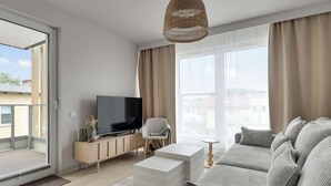 Ein modernes Wohnzimmer mit einer Couch, einem Flachbildfernseher, einer Weidenpendelleuchte und einem großen Fenster mit Vorhängen.