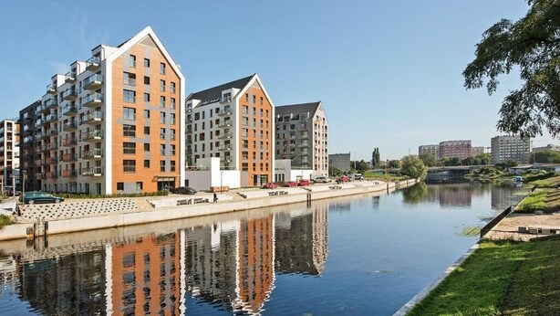 Un immeuble d’appartements moderne, doté de balcons, situé en bordure d’un canal et offrant une vue sur un ciel dégagé.