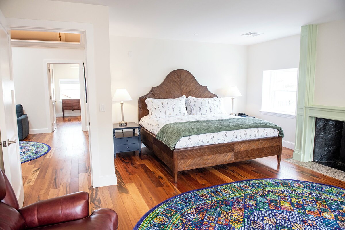 A bedroom with a wooden bed, two bedside tables, a chair, a patterned rug, and a fireplace.
