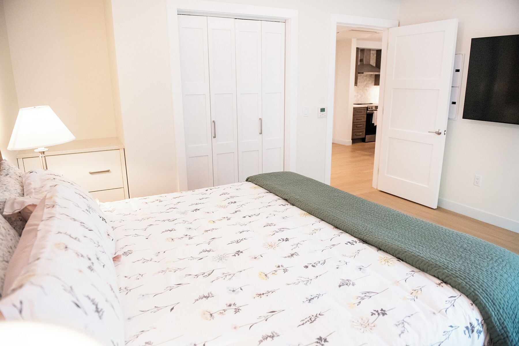 A bedroom with a bed, a nightstand, a lamp, a wardrobe, and an open door to a kitchen.