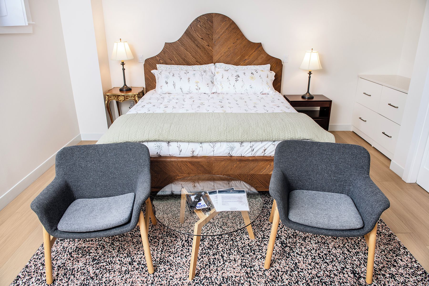 A bedroom with a wooden headboard, two bedside tables with lamps, a bed with floral bedding, a small table with a newspaper, and two grey armchairs.