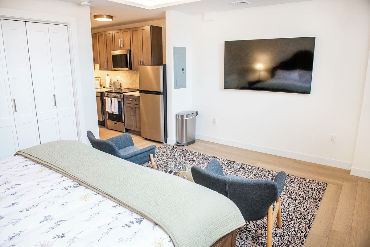 A modern bedroom with a bed, two chairs, a small kitchen area, and a TV mounted on the wall.