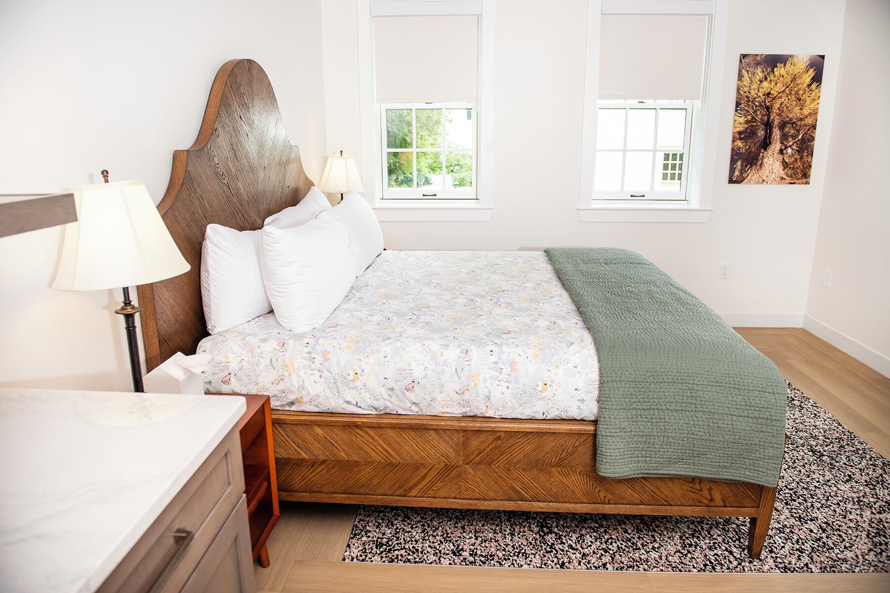 Un dormitorio con cabecera de madera, una cama con colcha estampada, un plaid verde, una mesita de noche con lámpara y una ventana con vistas al follaje.