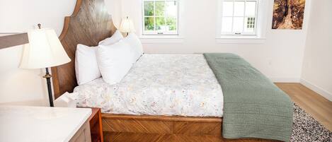 Un dormitorio con cabecera de madera, una cama con colcha estampada, un plaid verde, una mesita de noche con lámpara y una ventana con vistas al follaje.
