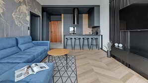 Ein modernes Wohnzimmer mit einem blauen Sofa, einem kleinen runden Tisch und einer Zeitschrift. Es gibt eine Küchenzeile mit einem Barhocker und einem Fernseher.
