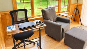 A room with a desk, a chair, a sofa, a lamp, and a view of a garden through large windows.