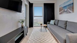 Ein modernes Wohnzimmer mit einem großen Flachbildfernseher, einem grauen Sofa, einem kleinen runden Tisch und Blick durch ein großes Fenster.