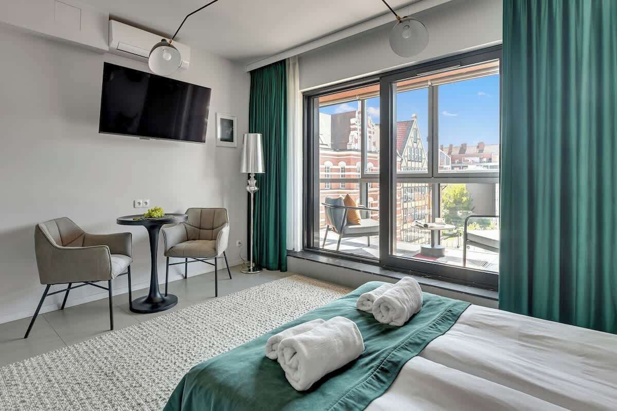 Habitación de hotel con cama, dos sillas, una mesita, televisión y un ventanal con cortinas.