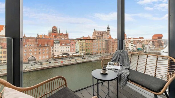 Ein Balkon mit Korbmöbeln, einem Tisch mit Tasse und Blick auf einen Fluss sowie historische Gebäude.