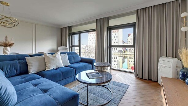 A modern living room with a blue sofa, a round coffee table, and a view of the city through large windows. - Rezydencja Wintera Grande | River View (Gdańsk)