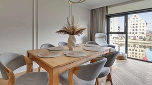 A modern dining area with a wooden table, grey chairs, a vase with dried plants, and a view of a cityscape through large windows. - Rezydencja Wintera Grande | River View (Gdańsk)